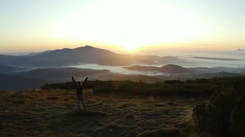 A hiker standing on a rock and raising hands in a gesture of victory on top of a mountain during sun