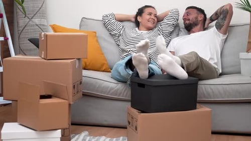 Young Couple Leaning on Sofa Surrounded By Unpacked Boxes Real Estate Journey to Their New Home
