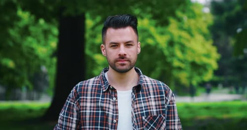 Slow motion of young man is looking and smiling in camera on green city park background in a sunny