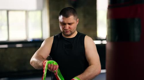 in a Boxing Fight Club Senior Muscular Trainer in a Black Tshirt Wraps an Elastic Multicolored