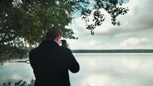 A Guy in Black Glasses Takes Pictures on His Phone on the Shore of the Lake