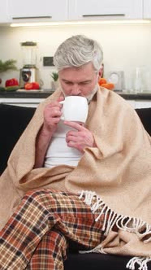 Man with blanket drinks tea on couch indoors