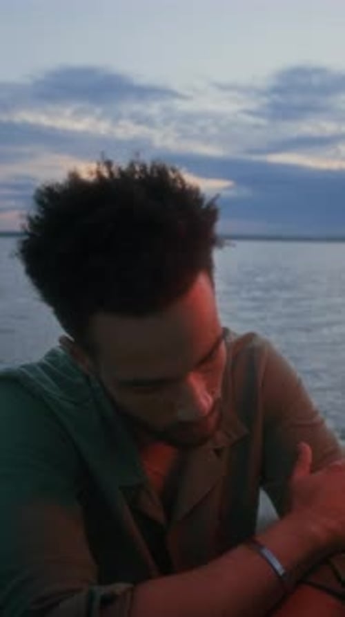 Vertical of Depressed Young Man Sitting on Sea Shore in Evening