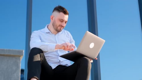 A Handsome Man is Sitting Outside Near an Office Center and Working on a Laptop Businessman is