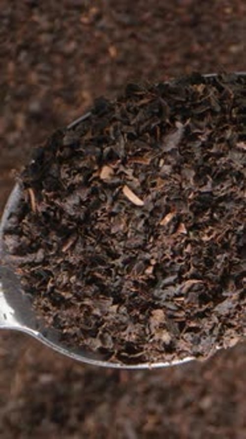 Vertical video. Ground dry black tea leaves on a metal spoon viewed from above against a rotating.
