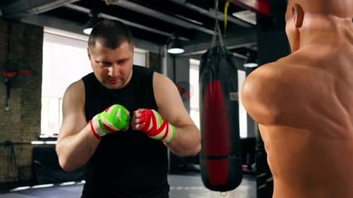in a Boxing Fight Club Senior Muscular Trainer in a Black Tshirt and with Multicolored Bandages is