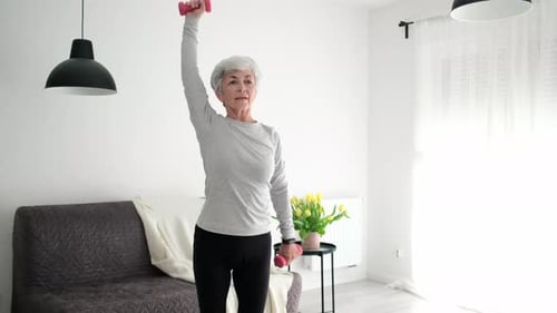 Atractiva mujer mayor de pelo gris haciendo ejercicios de fitness con mancuernas en casa