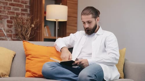 Focused Male Hipster Turn Pages Reading Book on Cozy Couch
