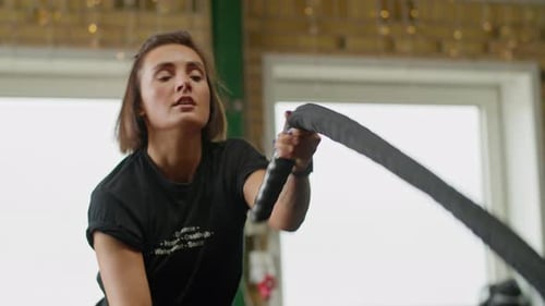 Active motivated female athlete training in gym, having a crossfit session with battle ropes, achiev