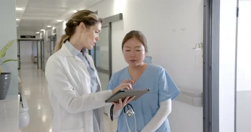 Focused diverse female doctors using tablet discussing in hospital corridor, slow motion