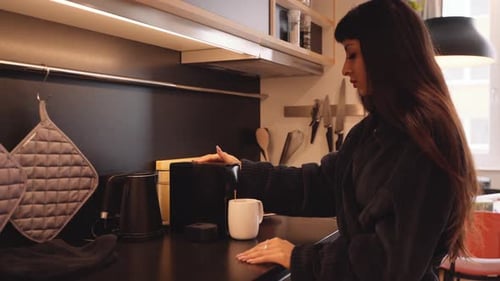 Woman in comfy robe in kitchen uses to make cup of coffee, profile view