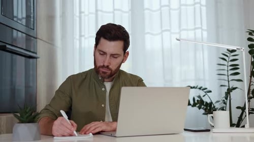 Man Working on Laptop and Writing at Desk