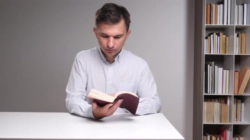 Man Focusing on a Book While Sitting at a White Desk with a Bookshelf Filled with Various Titles in