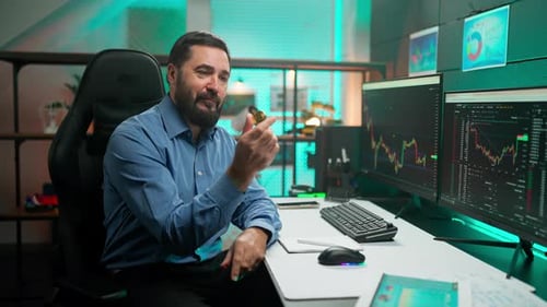 Man Holds Gold Coin at Computer Desk