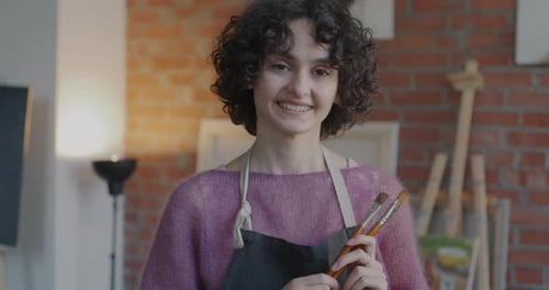 Slow Motion Portrait of Cheerful Young Lady Artist Wearing Apron Standing in Workshop with Pictures