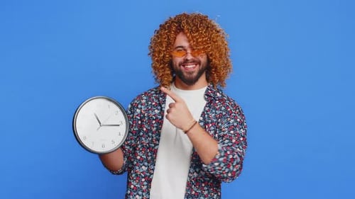 Aufgeregter kaukasischer Mann zeigt die Uhrzeit auf der Wanduhr im Büro an. Ok, Daumen hoch, Genehmigen, Beeilen Sie sich, Frist