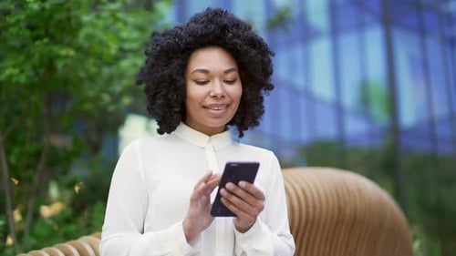 Young african american female employee is using smartphone sitting on a bench on street near