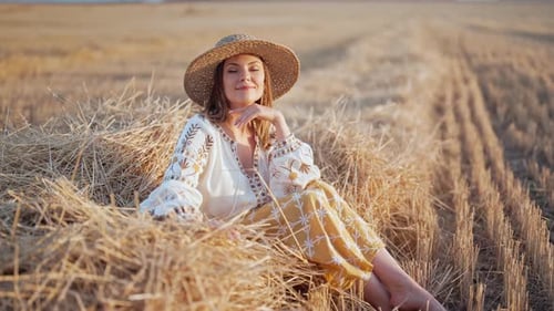 Pretty Woman in Straw Hat and Embroidered Blouse Smiling Lying on Hay in Countryside at Sunset
