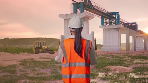 Asian Female Looking At Blueprint In His Hands While Walking at Construction Site