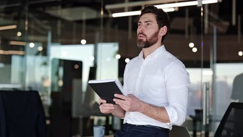 Bearded Businessman in White Shirt Writes in Notebook and Smiles in Modern Office