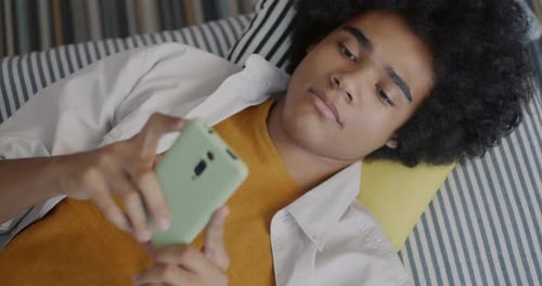 Top View of African American Teenage Kid Using Smartphone Lying in Bed at Home