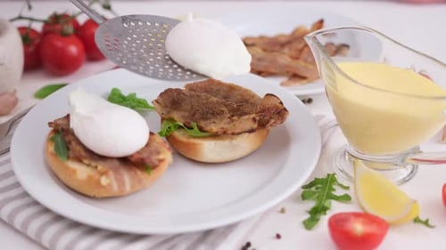 Making Egg Benedict Woman Puts Poached Egg on a Grilled Bun with Fried Bacon and Arugula
