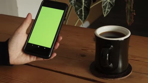 Female hand sitting on table drinking coffee and swiping up on green screen of smartphone