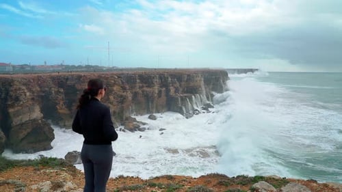 Woman in Black Sportswear Stands on a Rocky Cliff Watching Massive Waves Crash Against the Rugged