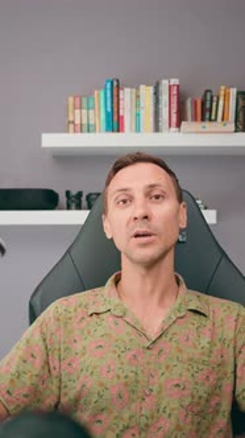 A Man is Seated in Front of a Bookshelf Surrounded By Books and a Floral Shirt in the Background