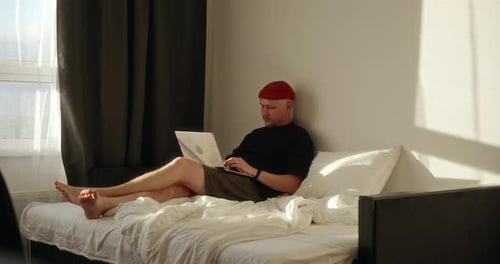 A Male Freelancer Works From Home Sitting on a Bed Using His Laptop