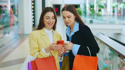 Two Happy Friends Stand in the Shopping Center are Looking at Smartphone Discussing Their Recent