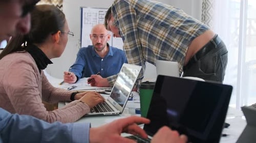 White Female Analyst Typing on Laptop While Team Leader and Colleagues Lean Over Table Reviewing