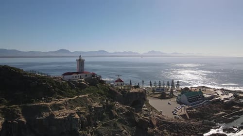 Drone shot flying over mountain cliff towards a lighthouse with ocean in the background on a sunny d