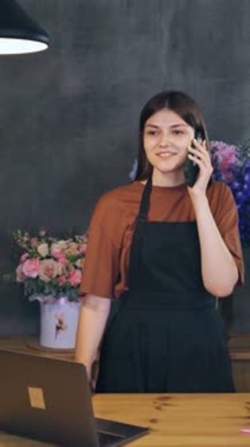 Woman Using Laptop and Phone in Flower Shop Surrounded By Plants and Flowers A Call From a Seller to