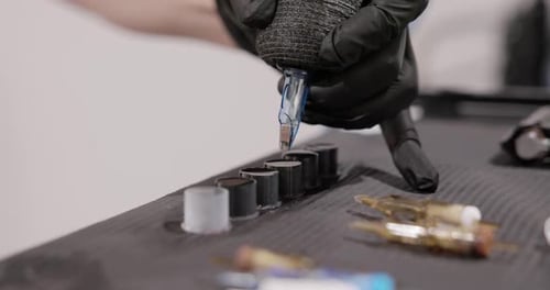 Tattoo Artist Dips The Tip Of The Needle Cartridge Into A Filled Ink Cap On The Table. - close up sh