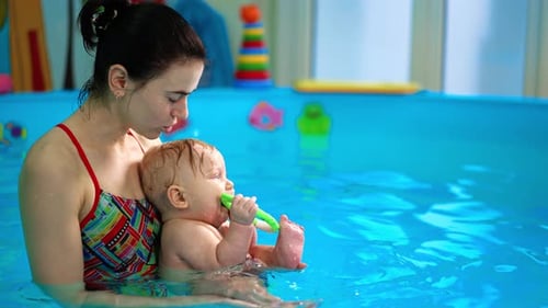 Woman with a little baby boy in the swimming pool. Adorable infant holds the toy in his mouth.