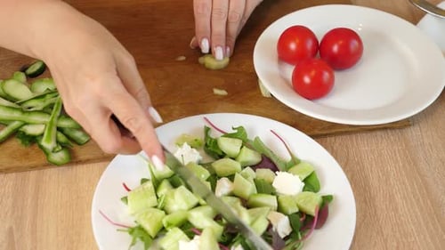 20 1Frau schneidet frische rote Tomaten auf einem Schneidebrett und legt sie auf einen Salatteller mit Gurken,