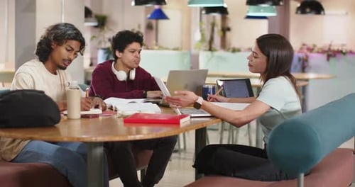 Three students in campus lounge, woman showing paper sparking study while center typing laptop