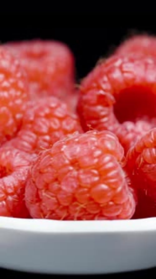 Vertical video. A raspberry in a small white plate, rotating in a loop on a black background.