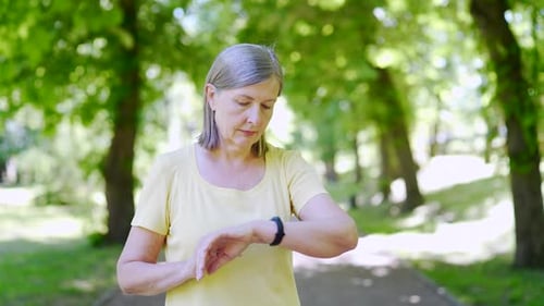 Mature female runner uses bracelet fitness tracker. senior gray-haired retired woman in summer park