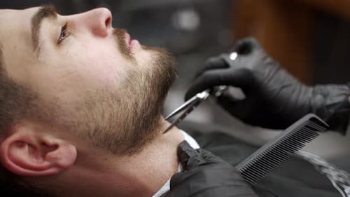 Barber Shapes Beard with Scissors on Young Man in Salon Grooming Stylist Works on Facial Hair Trim