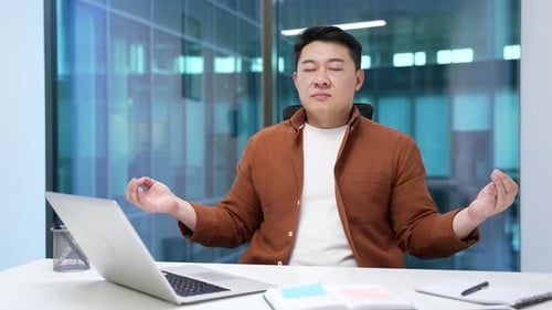 Asian businessman meditating at workplace in modern office with closed eyes and relaxed expression.