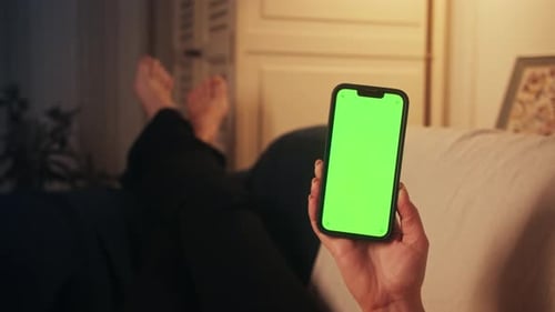 Woman holding a smartphone with a green screen. Woman relaxing on a sofa at home.
