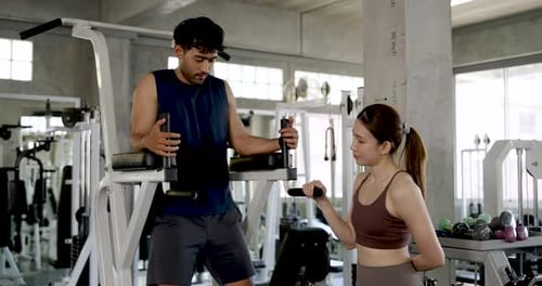 Young Asian couple preparing partner workout in modern gym, man explains knee raise machine to