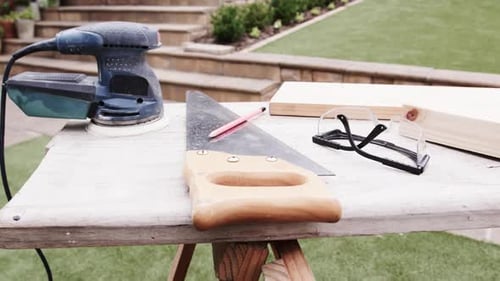 Backyard bench showing saw with wooden handle resting diagonally with red pencil, sander, glasses