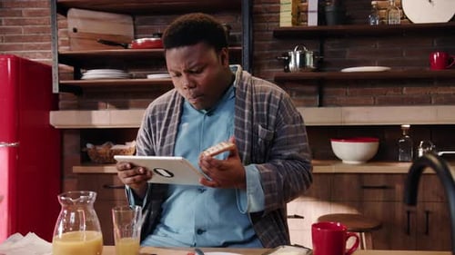 A Cheerful Man in His Cozy Kitchen Uses a Tablet While Enjoying a Delicious Breakfast