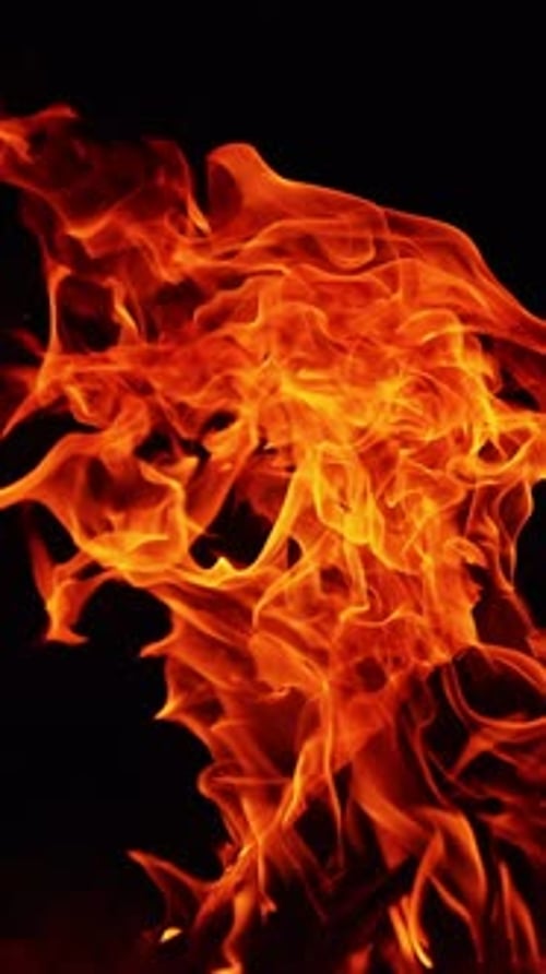 Auf
einem Haufen gestapelte Holzscheite brennen in einem großen Lagerfeuer. Feuerflammen biegen sich in der Dunkelheit nach rechts.