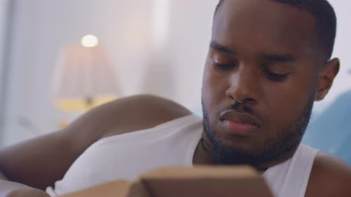 Young Man Relaxing Lying on Bed and Reading a Book in Bright