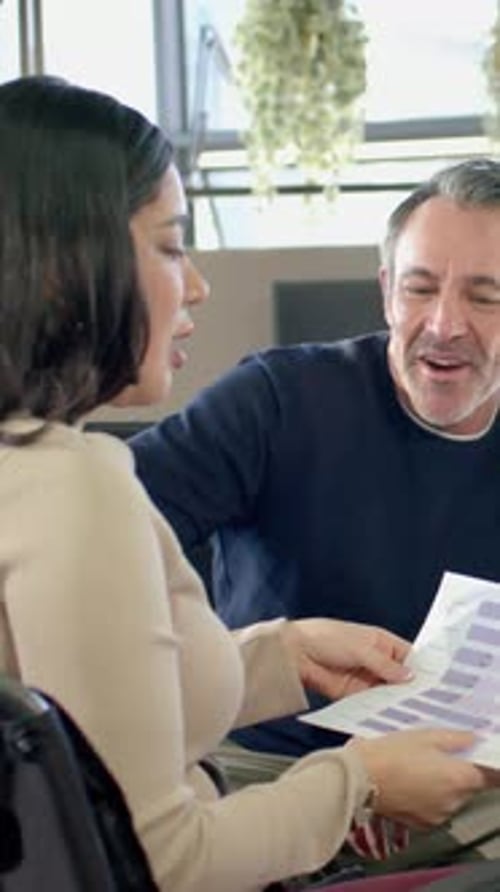 Vertical video: Holding chart, woman beige top, man navy sweater working at office reviewing charts
