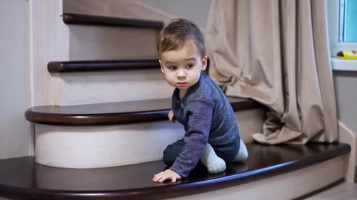 Cute little boy goes down by the stairs indoors. Toddler moves backward carefully watching his way.